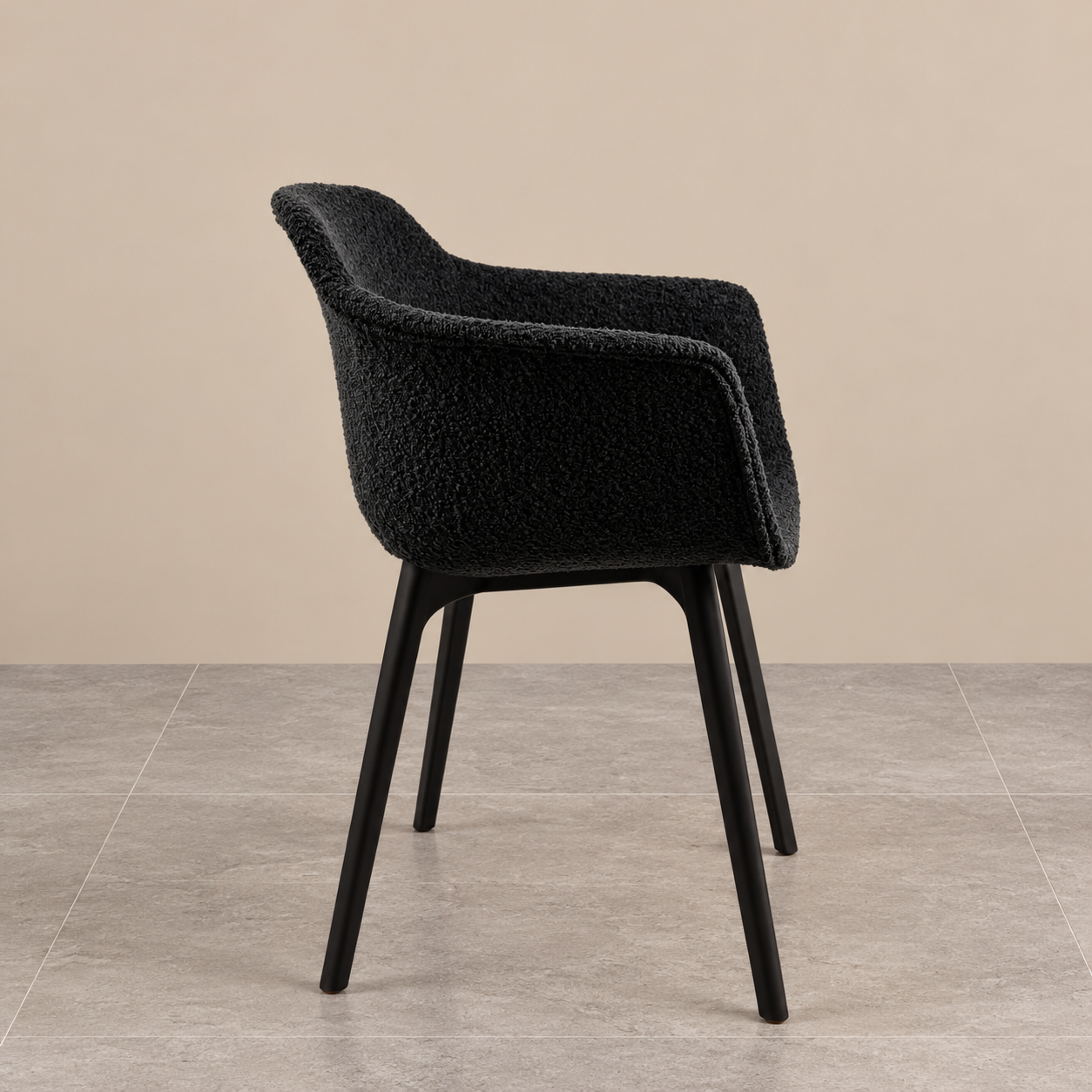 Side view of a contemporary black bouclé dining chair featuring textured upholstery, a sculptural curved backrest, integrated armrests and slim matt black legs, photographed against a warm beige background and grey tiled floor.