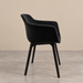 Side view of a contemporary black bouclé dining chair featuring textured upholstery, a sculptural curved backrest, integrated armrests and slim matt black legs, photographed against a warm beige background and grey tiled floor.