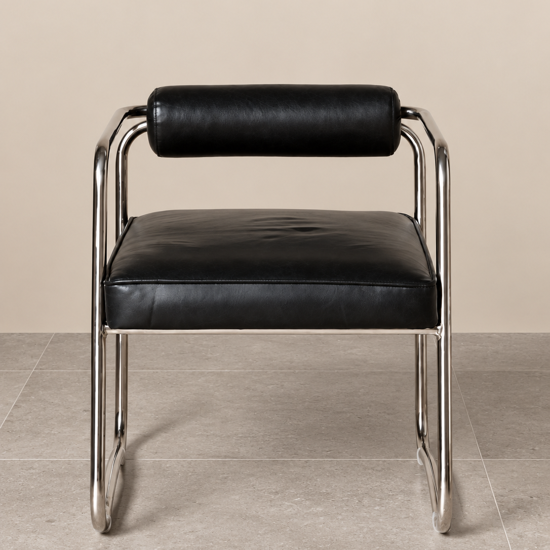 Front view of a retro-inspired accent chair featuring soft black leather upholstery, a cushioned cylindrical backrest and sculptural matt chrome tubular frame, against a warm beige wall and grey tiled floor.