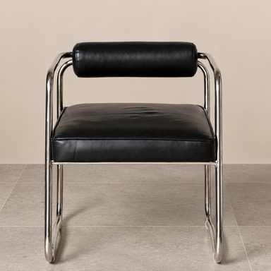 Front view of a retro-inspired accent chair featuring soft black leather upholstery, a cushioned cylindrical backrest and sculptural matt chrome tubular frame, against a warm beige wall and grey tiled floor.