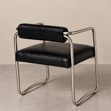 Retro-inspired accent chair featuring soft black leather upholstery, a cylindrical backrest and sculptural matt chrome tubular frame, photographed against a beige wall and grey tiled floor.