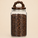 Glass storage jar filled with coffee beans, featuring a brown ceramic braided-handle lid against a warm beige background.