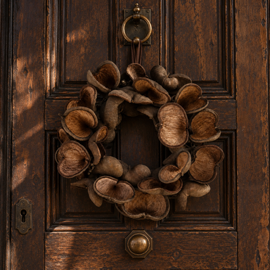 Natural brown dried botanical wreath featuring sculptural seed pods in warm walnut and chocolate tones, hanging on an antique dark wooden exterior door.