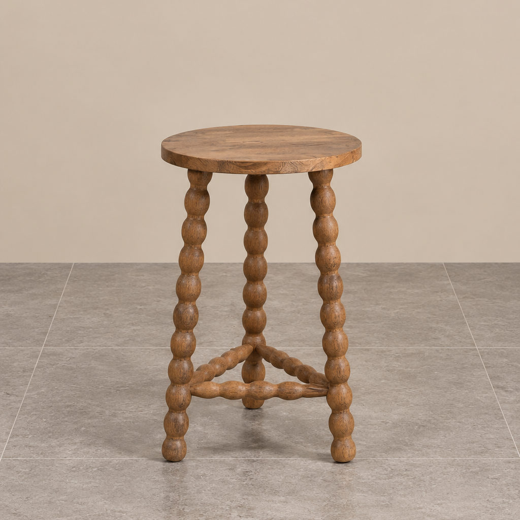 Natural wooden bobbin side table featuring a round tabletop, decorative beaded legs and sculptural wooden supports, photographed against a warm beige wall and grey tiled floor.