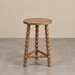 Natural wooden bobbin side table featuring a round tabletop, decorative beaded legs and sculptural wooden supports, photographed against a warm beige wall and grey tiled floor.