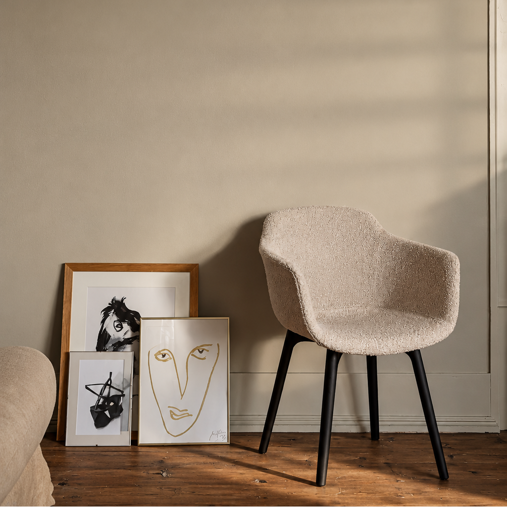 Cream bouclé sculptural dining chair with curved armrests and slim matt black legs, styled in a contemporary interior with neutral walls, wooden flooring and framed artwork.