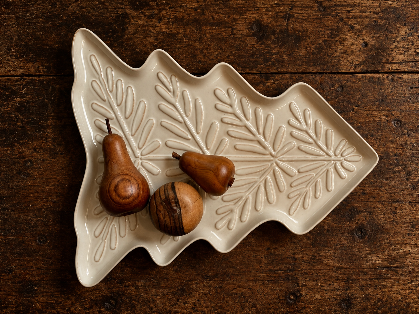 Ivory ceramic Christmas tree-shaped platter featuring raised foliage detailing and a glossy finish, styled with three decorative wooden ornaments on a dark vintage oak dining table.