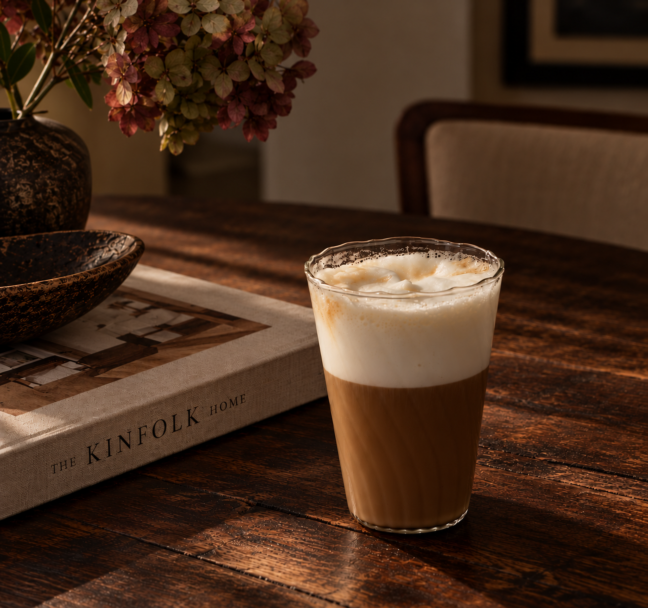 Clear ripple glass filled with a frothy latte, styled on a dark walnut table beside an interior design book and decorative flowers.