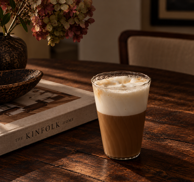 Clear ripple glass filled with a frothy latte, styled on a dark walnut table beside an interior design book and decorative flowers.