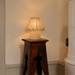 Natural beige pleated fabric table lamp with a decorative wavy shade, illuminated on a vintage dark wooden stool against a warm neutral wall.

