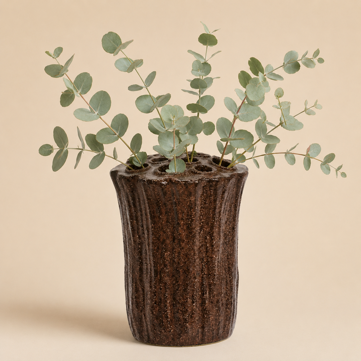Sculptural brown tree trunk vase styled with eucalyptus stems against a warm beige background.