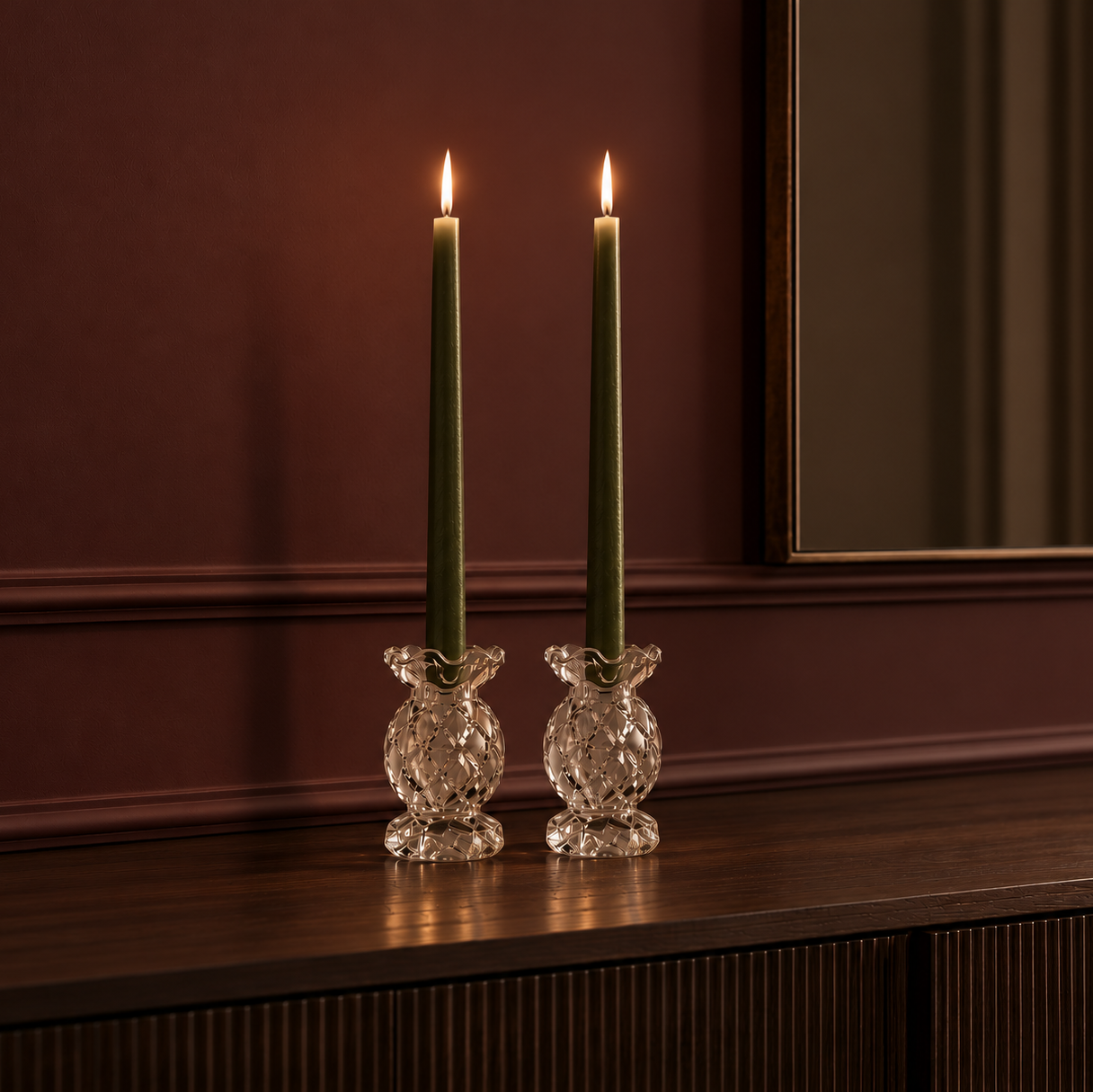 Two vintage-inspired clear cut glass candle holders with tall olive green dinner candles, styled on a dark walnut sideboard against a rich burgundy wall.