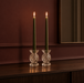Two vintage-inspired clear cut glass candle holders with tall olive green dinner candles, styled on a dark walnut sideboard against a rich burgundy wall.